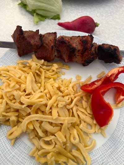 A close-up, eye-level shot shows a meal on a plate with a light blue patterned rim. A skewer of grilled meat, appearing to be pork or beef, is positioned horizontally across the top portion of the plate, with some pieces showing char marks. To the left and bottom of the skewer is a pile of spaetzle, a type of egg noodle, which are golden yellow and appear to be freshly cooked. To the right of the spaetzle, two slices of red bell pepper are placed on the plate. In the background, out of focus, is a piece of lettuce and a single red radish. The lighting suggests an outdoor setting, possibly a backyard or patio. The scene is devoid of any people or animals. The location is identified as Schwielowsee, Germany.