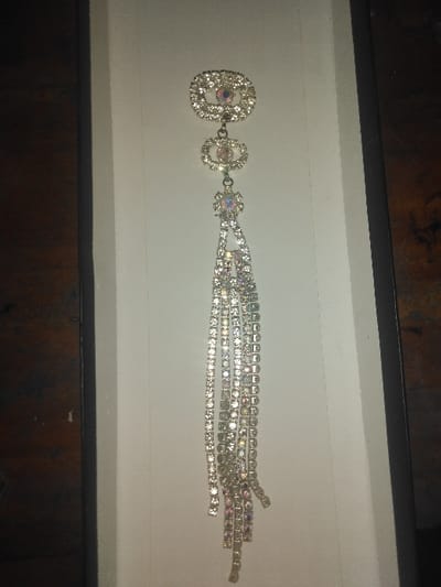 The image displays a sparkling ornamental brooch or pendant, approximately 15-20 cm in length, situated within a white-lined rectangular container or box. The object is constructed from numerous faceted clear and iridescent rhinestones or crystals set in a metallic, likely silver-toned, base. The design features three connected oval-to-triangular shaped segments at the top, each embellished with smaller surrounding stones and a larger central iridescent stone. From the lowest segment, multiple individual strands of square-cut stones cascade downwards, forming a tassel-like effect. The strands vary slightly in length, creating an uneven bottom edge. The white container is framed on its left and right by a dark brown, textured surface, possibly wood grain. The scene is illuminated by artificial light, causing reflections and showcasing the multi-spectral iridescence of the stones. No individuals are present. This scene is located in Gerasimovskoye, Kazakhstan.