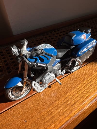 This media file features a close-up, angled shot of a toy motorcycle, a replica of a Carabinieri police motorcycle, resting on a textured wooden surface. The motorcycle is predominantly blue and silver, with prominent "CARABINIERI" text in white lettering on the blue bodywork. A red and white emblem, likely a police insignia, is visible on the left side of the motorcycle. The detail of the toy is high, showing components like wheels, handlebars, suspension, and exhaust pipes.

The wooden surface appears to be part of a piece of furniture or a decorative element, with a clear wood grain texture. The lighting suggests it might be daytime, with sunlight casting shadows and highlighting the details of the motorcycle and the wood. The background is out of focus, with a dark, textured area to the left and the wooden surface dominating the right. The setting appears to be indoors, given the texture and appearance of the background elements. No people are visible in the frame. The overall impression is one of a detailed collectible item presented in a focused shot.
