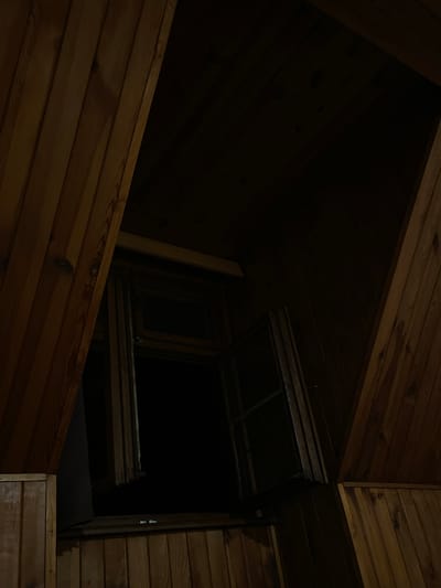 The image depicts the interior of a dimly lit, wood-paneled room, possibly an attic or a cabin. The focus is on an open window with dark panes, framed by wooden beams and planks. The wooden textures are prominent, with visible grain and knots. The overall impression is one of shadow and rustic simplicity. No people or specific landmarks of Jihlava, Czechia are visible.
