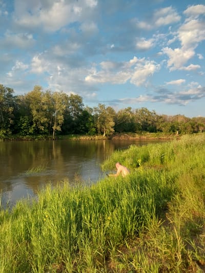 A man is crouched by the edge of a calm river, his back to the camera. He is partially submerged in the water, with only his upper body and head visible. The riverbank is covered in tall, green grass, some of which is bent over where the man is sitting. Trees line the opposite bank of the river, creating a dense, green backdrop. The sky above is blue with scattered white clouds. The scene appears to be a natural setting, possibly in Chkalov, Russia, given the context.