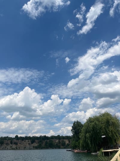 The image depicts a picturesque scene at a body of water, possibly a quarry or a lake, in Novomykolaivka, Ukraine. The sky is a vibrant blue, dotted with fluffy white cumulus clouds. A steep, rocky cliff covered with lush green trees forms the backdrop on the far side of the water. On the right side of the frame, a large, weeping willow tree dominates the scene, its branches cascading downwards towards the water. Beneath the willow, there is a wooden pier with several people lounging on inflatable mats, enjoying the sunny day. Another smaller wooden structure is also visible. The water is calm and reflects the blue sky. Several boats are moored along the pier. The overall impression is one of a tranquil and idyllic summer day in a natural setting.