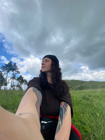 A medium shot from a low angle shows a woman with long, dark, wavy hair and tattoos on her arms, wearing a black beret and a dark brown t-shirt. She is outdoors, in a grassy field with rolling green hills in the background and pine trees to her left. The sky is overcast with patches of blue visible. The woman is looking to her right, with her left arm extended forward, holding what appears to be a phone or camera. A silver bracelet is visible on her left wrist. The overall scene suggests a natural, possibly rural, outdoor setting. There is no indication of this location being in Andreevka, Russia.