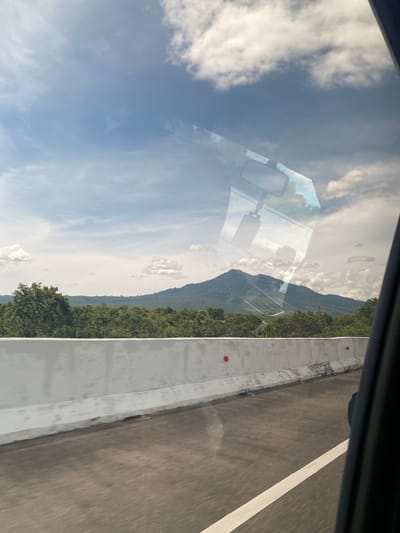 A view from inside a moving vehicle shows a section of a highway in Seulimeum, Indonesia. The highway is bordered by a concrete barrier with intermittent red markers. Beyond the barrier, dense green vegetation climbs a hill. The sky is partially obscured by clouds and the vehicle's window, which reflects interior elements like the rearview mirror. The road surface appears dark gray with a white lane marker.