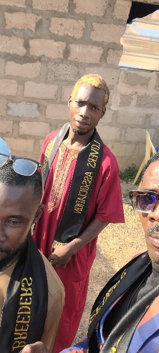 A close-up, outdoor photograph depicts three adult males, likely in a selfie-style shot, against the backdrop of an unfinished brick wall in the city of Alayi, Nigeria. The central figure, an individual with dark skin and blonde-dyed hair, wears a red traditional tunic with gold embroidery and a black sash bearing the inverted gold text "LOVERS ASSOCIATION." He faces the camera directly with a neutral expression. To his immediate left, partially visible in the foreground, is another male with dark skin, wearing sunglasses on his head and a tan garment, also with a black sash displaying inverted gold text, partially legible as "BREEDERS." To the central figure's right, partially visible in the foreground, is a third male with dark skin, wearing dark sunglasses, a mustache, and a blue garment, also with a black sash. The background features a light-colored concrete block wall with visible mortar and a small patch of dry ground vegetation. A piece of corrugated metal is visible in the upper right background. The scene is illuminated by bright daylight.
