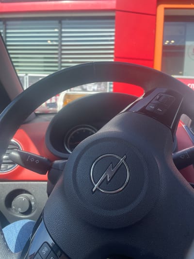 The image shows the steering wheel of an Opel car, with the Opel logo prominently displayed. The car is likely parked in front of a building with a red exterior. Reflections in the windshield suggest a bright day and a cityscape or built environment outside. The image does not provide any information to confirm it is in Vodenichane, Bulgaria.