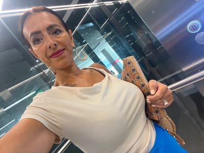 A medium-shot, selfie-style photograph captures a woman in an elevator. She is positioned in the lower-left quadrant of the frame, her face angled towards the camera, and her body is slightly turned to the right. The woman has medium-toned skin, dark hair pulled back, and is wearing dark eyeliner and pink lipstick. She has large hoop earrings, one of which is visible on her left ear. Her attire consists of a white, ribbed, short-sleeved top and bright blue pants. A patterned clutch bag is held in her right hand, which rests on her left side. A silver ring is visible on her right index finger. A tattoo is visible on her left shoulder blade. The elevator's interior is characterized by reflective glass walls, a stainless steel panel with illuminated elevator buttons, and fluorescent lighting. The backdrop suggests an urban environment, possibly within a modern building. The location is identified as the city of Mensdorf, Luxembourg.