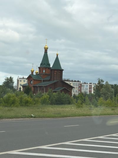 The image shows a wooden Orthodox church with green roofs and golden domes, located in the city of Kstovo, Russia. The church is surrounded by lush green trees and foliage. In the background, there are modern apartment buildings. The foreground features a paved road with white lane markings, suggesting a view from a vehicle. The sky is overcast with clouds. There are no people or animals visible in the scene.