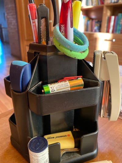 This is a close-up, indoor photograph of a desk organizer filled with stationery items. The organizer is black and appears to be made of plastic. Prominently featured are a yellow highlighter, red and white pens, and a pair of colorful hair ties (one blue, one green). A silver stapler is positioned above and behind the organizer.
Inside the organizer, a single AA battery is visible, with warning text indicating "ZERO MERCURY", "WARNING: May explode or leak if recharged, disposed of in fire or dissected. Made in China." Next to the battery, a portion of a yellow highlighter is visible, with the text "STABILO BOSS MINI" clearly readable. Another cylindrical object, possibly a container of some sort, with blue and white labeling and blue cap, is also present.
The background shows a wooden bookshelf filled with books, suggesting a home or office setting. The lighting appears to be natural, possibly from a window, creating some glare and casting shadows. The overall scene conveys a sense of organization and preparedness, with various writing and office supplies readily accessible. There are no people or animals visible in the image, nor any specific indication of the time of day or weather conditions. The setting appears to be tidy, though some dust is visible on the organizer.