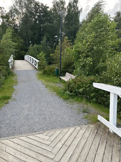 The image depicts a gravel path leading to a white arched bridge, which spans over a small body of water that is not fully visible. The bridge has a wooden walkway and white railings. To the right of the path, there is a green trash can and a wooden park bench nestled amongst dense green foliage. A lamppost is visible behind the bench, and further to the right, a section of a white railing is prominent in the foreground. The surroundings are lush with mature trees and undergrowth, suggesting a natural or park-like setting. The sky above is overcast with some light filtering through. The scene appears to be an outdoor public space, likely a park or walking trail, within the city of Vammala, Finland, known for its scenic natural beauty.