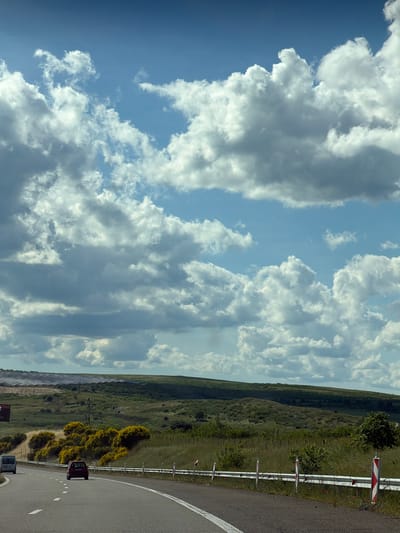 The image depicts a road winding through a landscape near Vratitsa, Bulgaria. The sky is partly cloudy with white cumulus clouds. The road has a white line marking the edge and safety barriers. The terrain consists of green, hilly meadows. In the background, solar panels are visible. A red and a white car are on the road.