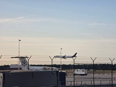 The image shows an airport scene at dusk, with clear skies and a few wispy clouds. In the foreground, a chain-link fence topped with barbed wire runs horizontally across the frame, partially obscuring the view of the tarmac. To the left, a portion of a large, gray container is visible. Beyond the fence, two aircraft are present. The tail of a white airplane with a black snowflake-like logo is visible on the left, with the registration "D-NICE" on its fuselage. Further to the right, a white Lufthansa airplane is in the air, appearing to be either landing or taking off, with its landing gear extended. Several traffic cones are scattered on the tarmac. In the background, a line of trees borders the airport. The overall impression is that of an active but quiet airport environment. This image was taken at Frankfurt Airport, located in Kelsterbach, Germany.