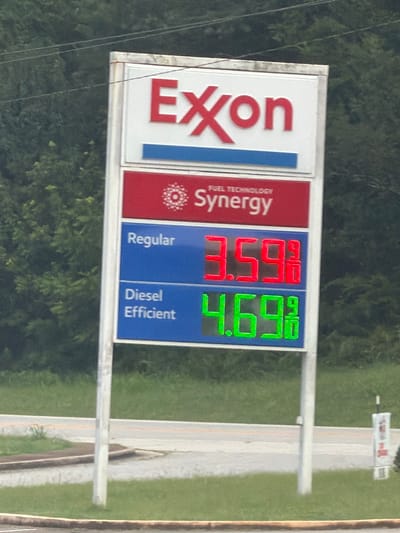 An outdoor gas station sign is visible in Wildwood, United States. The sign, mounted on a white pole, displays the Exxon logo at the top in red lettering. Below the logo, "FUEL TECHNOLOGY Synergy" is written on a red background. The sign then shows prices for different types of fuel. "Regular" fuel is priced at 3.59 9/10 dollars per gallon, displayed in red digital numbers. "Diesel Efficient" fuel is priced at 4.69 9/10 dollars per gallon, displayed in green digital numbers. The background consists of lush green trees and foliage. The sign is positioned in front of a grassy area with a road visible in the lower portion of the image.