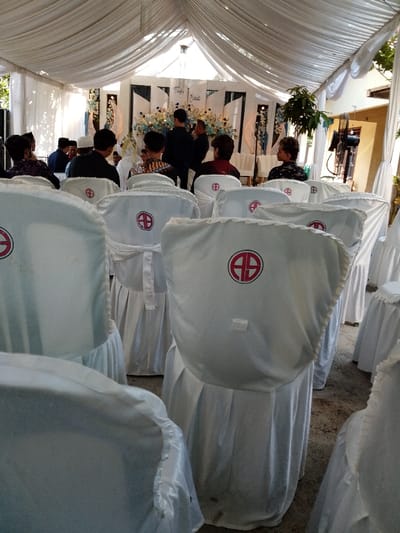 The image depicts an indoor/outdoor event, identified as a wedding ceremony for 'Taufiq & Rizka,' located in Juwana, Indonesia. The scene is illuminated by daylight filtered through a white fabric tent canopy. Numerous chairs are arranged in rows, covered with white fabric slipcovers that extend to the floor. Each visible chair cover prominently displays a red circular emblem containing the white letters "AB." The flooring beneath the chairs is a hard, discolored surface, likely concrete.
In the background, a decorated stage area features white panels adorned with light blue and white floral arrangements, along with crescent moon-shaped decorative elements. The names "Taufiq & Rizka" are visible on a central panel. Guests are seated in the foreground and middle ground, primarily with their backs to the camera, observing the stage. They are dressed in semi-formal attire, including dark shirts and traditional head coverings. Several individuals are standing near the stage, some in dark clothing, with one person partially visible in white attire. To the right of the seating area, a large, dark-colored fan or lighting stand and a small potted plant are visible.