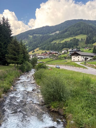 The image shows a scenic view of a village nestled in a mountainous valley. The village consists of several buildings with traditional architecture, featuring pitched roofs and multiple stories. A paved road winds through the village, with a few cars visible. The surrounding landscape is characterized by lush green hillsides covered in dense coniferous forests, ascending to higher, mist-covered peaks. A clear, fast-flowing stream or river runs alongside the road and through the foreground, with white water churning over rocks. The vegetation is vibrant green, with a mix of trees and grass, indicating a temperate climate. The sky is partly cloudy, with large, fluffy white clouds against a bright blue backdrop. The overall scene evokes a sense of peacefulness and natural beauty. The orientation of the image suggests it was taken with the camera held vertically, likely to capture the expansive view of the valley and mountains. The time of day appears to be daytime, given the bright sunlight and visible shadows. The location appears to be Ratschings - Racines, Italy, as indicated by the context, suggesting a mountainous region in the Alps. There is no visible text in the image.