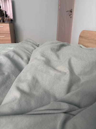 The image shows a close-up of a rumpled light gray blanket. In the background, there is a section of a wall painted a light blue color. A small portion of a light brown wooden cabinet or dresser is also visible. Additionally, a light pink or white headboard can be seen near the top left corner of the image, featuring a decorative design and visible hardware. The photo does not provide enough information to determine a specific location in Nadarevo, Bulgaria, or any information about people, interactions, or actions.
