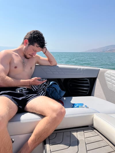 A young man is sitting in the rear of a boat, facing the stern. The boat is on a body of water with a distant mountain range in the background. The man is wearing a black swimsuit and is looking down at a phone in his hand. The boat has gray seats and a black speaker in the back. The man is sitting on a white, cushioned seat, and his legs are stretched out in front of him. There is a blue object, possibly a towel, to his right. The boat is docked on the Sea of Galilee, which is located at the coordinates provided. The sky is clear, and the sun is shining.