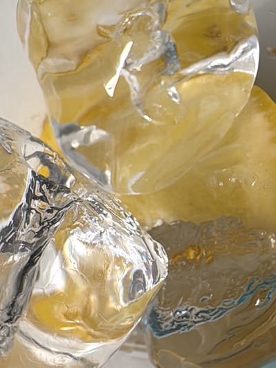 The image presents a close-up, abstract view of multiple translucent and transparent objects. These objects exhibit irregular, faceted shapes consistent with ice cubes, displaying clear, reflective surfaces with prominent specular highlights and refractions. Embedded within or behind these transparent structures are areas of a yellow-amber hue, suggesting either a colored liquid surrounding the ice or a material of that color. No discernible people, actions, or specific environmental details beyond the immediate subjects are present. The scene is located in Peschiera Borromeo, Italy.