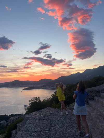 Two women stand on a stone surface overlooking a lake and a mountain range at sunset. The woman on the left wears a yellow dress. The woman on the right wears a blue shirt and white shorts and is looking at her phone. The sky is filled with pink-tinged clouds. Boats are visible on the lake, and a city stretches along the shore. The scene appears to be taken in Vojnići, Montenegro.