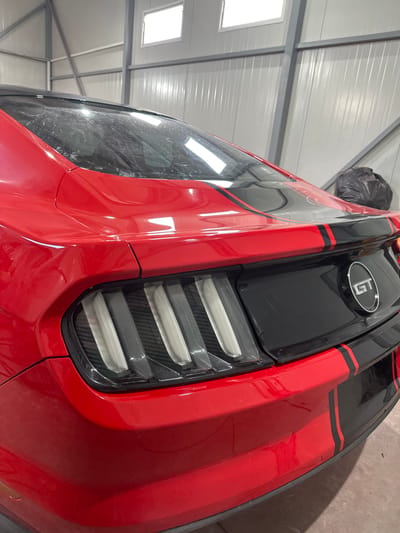 A close-up, angled view shows the rear of a red Ford Mustang GT parked inside a metal building. The taillights are horizontally striped and illuminate white. A black racing stripe with thin red borders runs vertically down the car's rear. A "GT" emblem is visible on the black portion of the car. The location is Golyanovtsi, Bulgaria, based on additional context not visible in the image. The area appears to be a garage or storage space.