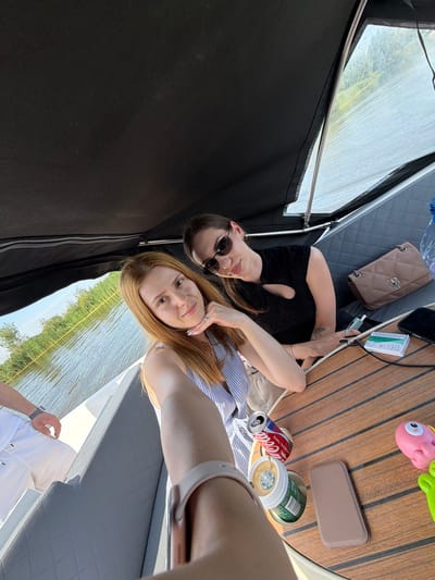 Two women are on a boat. The woman in the foreground is blonde and wearing a striped top. She is holding a Starbucks cup and a can of Coca-Cola. The woman in the background is wearing sunglasses and a black top. She has a purse on her lap. The boat is on a body of water with reeds on the shore. The scene appears to be a leisurely outing on a summer day. The location is Gellicum, Netherlands.