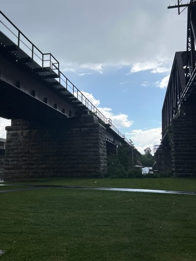 The image shows two large bridges constructed of metal and stone. The bridge on the left features a long, dark metal walkway with railings, supported by massive stone pillars. The bridge on the right is a more complex metal structure with visible trusses, also supported by stone foundations. Both bridges span over a body of water, with a line of trees visible in the background. The foreground consists of a well-maintained green lawn, with a paved pathway winding through it. The sky is cloudy with patches of blue sky, and there are visible rain droplets in the air. The scene appears to be an outdoor public space. This view is characteristic of the Sainte-Anne-de-Bellevue Canal, a waterway in Sainte-Anne-de-Bellevue, Quebec, Canada, which is spanned by several bridges, including railway bridges.