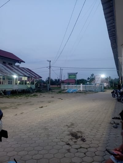 The image, rotated 90 degrees clockwise, captures a street scene at dusk or dawn, characterized by dim, soft light and a light blue-grey sky, possibly overcast. The setting appears to be a village or small town in Dayahtuha, Indonesia.
In the foreground, the paved ground is made of hexagonal interlocking pavers, showing some dark stains. On the far right, a person's arm and hand are visible, holding a smartphone. Part of a dark object, possibly a motorcycle, is also at the bottom right.
The midground features a street extending into the distance. To the left of the street, a utility pole with multiple power lines is prominent. Further down the street on the left, a building with a reddish-brown roof and light-colored walls is illuminated by bright lights, suggesting they are on or shining on the structure. A green sign with white text, partially visible below the utility pole, reads "DAYAH PESANTREN," indicating an Islamic boarding school.
Further along the street, another structure with a white fence and a bright light, likely a streetlamp, is visible. Trees and more distant buildings line the street, contributing to the serene, quiet atmosphere of early morning or evening. Another motorcycle is parked further down the road on the right.