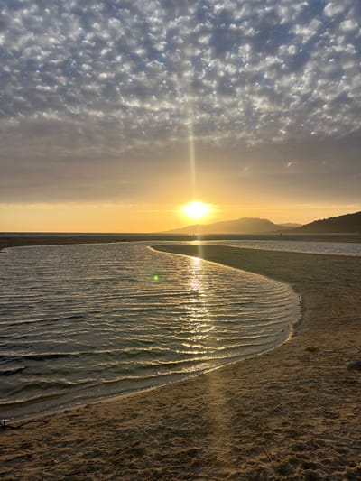 The image shows a scenic sunset at the beach in Tarifa, Spain. The sun, a bright yellow orb, is low on the horizon, casting a warm glow across the sky and reflecting on the rippling water. The sky is filled with scattered, textured clouds, creating a dramatic backdrop for the sunset. In the foreground, a sandy beach curves along the edge of the water, with gentle waves lapping the shore. In the distance, silhouetted mountains form a majestic outline against the bright sky. A few small figures can be seen on the beach and in the water, enjoying the serene atmosphere. The overall impression is one of natural beauty and tranquility.