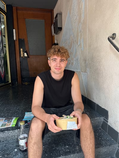 A young man with curly blonde hair and a black t-shirt is sitting on dark speckled steps outdoors. He is holding a small, light brown box with a label on it. His left arm rests on his lap, and his right arm is extended, holding the box. He is smiling at the camera. Beside him on the steps are a few items, including a partially visible bottle with a dark liquid and a label, a small stack of papers, and a green and white brochure. The background shows a wooden door with a glass pane, part of a wall with a decorative relief, and a metal handrail. The scene appears to be in the daytime, likely sunny given the lighting. The man's expression is relaxed and happy. The location context is Hamm, Germany. There is visible text on the label of the box and the bottle, but it is mostly illegible.