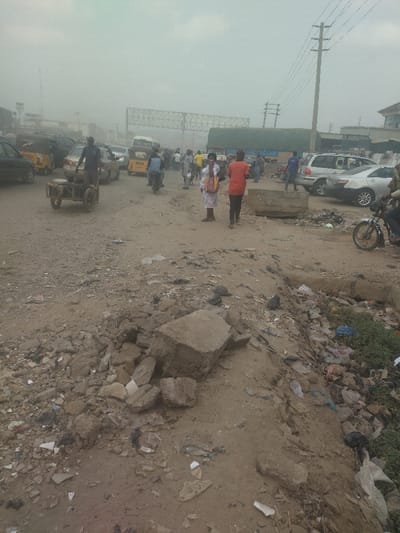 The image shows a street scene in the city of Way, Nigeria, at the coordinates 9.029 latitude and 7.584 longitude. The street is dusty and littered with debris, including broken concrete slabs, plastic bottles, and other trash. A man in a red shirt and a woman in a white and pink dress are walking down the street, and another man in a black shirt is pushing a handcart loaded with goods. There are several cars parked on the side of the road, as well as some pedestrians walking in the distance. The atmosphere is hazy and dusty, and the sky is obscured by a thick layer of haze. The image shows a clear lack of maintenance in the public space.