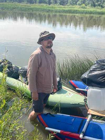 A man stands in shallow, muddy water next to several inflatable rafts loaded with black trash bags. He is wearing a camouflage hat, sunglasses, a light brown hoodie, and dark shorts. The rafts are green and blue. In the background, a calm river reflects the sky and a dense line of green trees. The scene appears to be a riverbank in a rural or natural setting, potentially near Nezhinka, Russia, given the context.