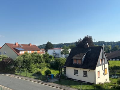 This is an exterior, eye-level, medium shot photograph of a residential neighborhood in Osterode am Harz, Germany. The sky is clear and blue. In the foreground, there is a paved road and sidewalk. Behind the sidewalk, there is a metal fence, a green lawn, and several houses. The house on the right is a two-story building with a dark, sloped roof and light-colored walls. It has several windows with orange shutters. A metal chimney extends from the roof. There is also a trampoline in the backyard of this house. To the left of this house is a green lawn with some bushes and trees. Further to the left, there are two other houses with red, sloped roofs. One of these houses has solar panels installed on the roof. The houses are surrounded by lush green trees and vegetation. In the background, there is a distant treeline and a clear blue sky. The image appears to have been taken on a sunny day.
