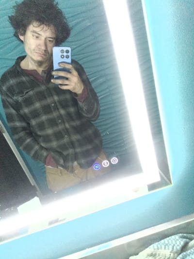 A man with dark curly hair takes a selfie in a bathroom mirror. He's wearing a plaid shirt over a maroon top and khaki pants. The mirror is illuminated with a bright LED light strip. The background features textured teal walls. The camera is a light blue smartphone with multiple lenses. Several control buttons are visible on the mirror's surface.
