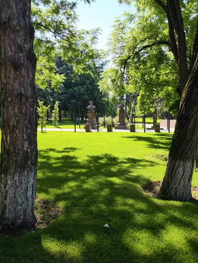 The image depicts a well-maintained park with lush green grass and mature trees. In the background, two statues or monuments are visible, each with a bust on a pedestal. The statues are located in a grassy area with lampposts and some decorative bushes or flowers. The overall impression is one of a peaceful and well-kept public space, likely within a city. The mention of Kolosisty, Russia, suggests this is a specific location within that city, though the image itself does not provide direct evidence of this. The scene is bathed in natural sunlight, casting dappled shadows on the grass from the surrounding trees. There are no people visible in the image.