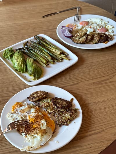 This is a top-down, medium shot image of two white plates of food on a light brown, wooden table. The plate in the foreground contains fried eggs, some with runny yolks, and a pile of fritters. A fork is on the plate, touching one of the eggs. The plate in the background features fried eggs, some with visible red yolks, a wedge of red onion, and a stack of fritters. A fork and knife are also visible on this plate. Another white plate in the middleground holds sliced avocado and cooked asparagus. The table surface has a visible wood grain pattern and a slight sheen. There are no people visible in the image. The lighting appears natural, casting soft shadows on the table. The scene depicts a meal, likely breakfast, served on a wooden table. There is no information provided about the location of Bakovići, Montenegro.