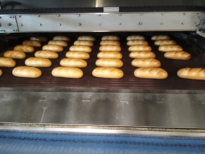 In the city of Șoldănești, Moldova, a conveyor belt oven is filled with rows of freshly baked bread. The bread, golden brown and uniformly shaped, are arranged in neat lines on a dark, textured surface. The oven itself is a large piece of industrial equipment, with metallic components and a visible conveyor system. The lighting suggests an indoor setting, likely a bakery or food production facility. No people are visible in the image, and the focus is solely on the bread and the baking machinery.