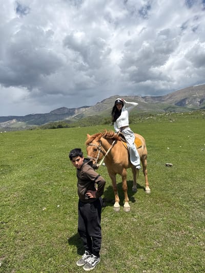 The image shows a woman lying across the back of a brown horse in a grassy, mountainous landscape near Teghut, Armenia. A man lies on the ground near the horse's front legs. The woman is wearing white clothing and a baseball cap. The man is wearing a dark jacket and pants with white sneakers. Mountains are visible in the background under a cloudy sky.