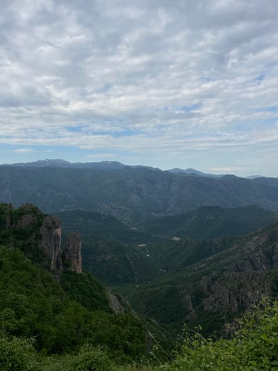 A scenic landscape captured in Kućišta, Montenegro, showcases a vast mountain range descending into a deep valley. The sky is filled with striated clouds. Lush greenery covers the foreground and parts of the mountain slopes. Rock formations are visible on the lower right, adding texture to the landscape.