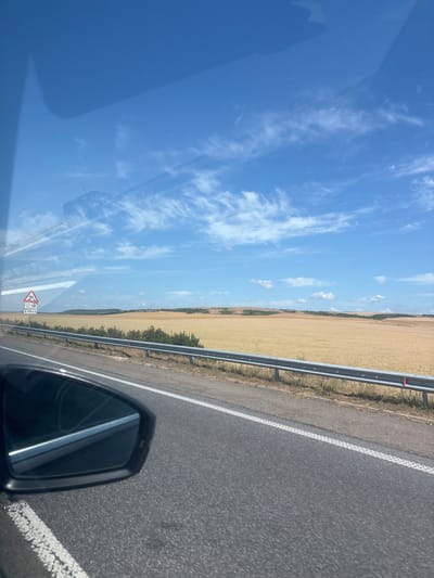 The image captures a view from a vehicle traveling along a paved road. In the foreground, the right side mirror of the vehicle is visible, reflecting a sliver of the road and sky. The road itself has white lane markings. To the right of the road, a metal guardrail separates the roadway from a vast field of golden crops, likely wheat or barley. Beyond the field, rolling hills covered with vegetation form the horizon. The sky is a bright blue, with scattered wispy white clouds. A triangular road sign is visible on the left side of the road, featuring symbols of a vehicle skidding, snow, and a number '2000m'. The image appears to have been taken on a sunny day. The scene suggests a rural or semi-rural environment, possibly in the vicinity of Troyanovo, Bulgaria, given the typical agricultural landscape and road infrastructure found in such regions.