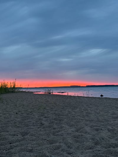 The image captures a serene sunset scene on a sandy beach. The sky displays a gradient of colors, transitioning from a soft blue at the top to a vibrant orange and pink band near the horizon, indicating sunset. The water of a lake or sea stretches across the middle of the frame, reflecting the colorful sky. The right side of the image is dominated by a sandy beach, with visible sand textures and a few blades of dry grass peeking through. The scene evokes a sense of peace and tranquility, typical of a quiet evening by the water. The time of day is clearly sunset due to the distinct sky colors. There is no visible text in the image. The location is identified as Володарский ауылдық округі, Kazakhstan.