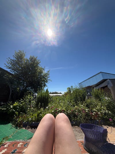 The image depicts a sunny outdoor scene in Karachino, Russia, with the sun prominently featured in the clear blue sky, creating a bright, lens-flare effect. The perspective is from ground level, looking upwards and slightly to the right.

The foreground is dominated by the legs of a person, suggesting they are sitting or reclining. Adjacent to their legs, a woven purple basket is visible, along with a patterned rug and a green mat. Behind these elements, a lush green garden or yard is in bloom with various plants and flowers in shades of pink and white.

In the background, a portion of a building with a corrugated metal roof is visible, along with what appears to be a stack of firewood. Another tree with dense foliage is also part of the landscape. The overall impression is of a relaxed summer day outdoors. The lighting indicates it is daytime, likely late morning or early afternoon, given the sun's intensity and position. There is no visible text in the image.