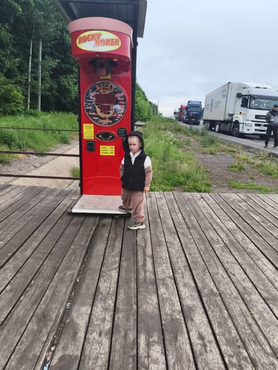 The image depicts an outdoor scene in Nizhnie Yaki, Russia, featuring a young child positioned on a weathered wooden plank platform. The child, a boy, wears a black hooded vest over a long-sleeved shirt with a white upper section and a light brown lower section, light brown pants, and dark sneakers with yellow accents. He stands with his hands in his pockets, looking forward with a serious expression, slightly to the right of a red "Rocky Boxer" arcade game.
The arcade machine, colored red, features a central punching bag mechanism and multiple display panels. The top section reads "ROCKY BOXER" in yellow lettering, while a lower circular display includes "ROCKY BOXING" and score categories such as "SUPER-MAN," "KILLER," and "CHAMPIONS." A vertical panel on the machine displays pricing in Russian rubles: "1 удар.....20р" (1 hit - 20 rubles), "2 удара.....50р" (2 hits - 50 rubles), and "5 ударов.....100р" (5 hits - 100 rubles). The machine rests on a white base on the wooden platform.
Behind the wooden platform, a narrow grassy embankment with a metal railing slopes down to a concrete channel. Further behind, a dense line of green trees and vegetation extends into the background. To the right of the scene, a paved road is visible, on which a queue of several large semi-trailer trucks is stopped or moving slowly. The closest truck is white with a large blue circular logo on its trailer, and a male figure in a gray and black jacket is standing near its front passenger side. Other trucks further down the road are blue and red. The sky above is overcast. The wooden platform itself shows signs of wear, with visible gaps between planks, some containing small pieces of debris.