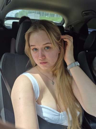 A young woman with blonde hair and blue eyes is sitting in the passenger seat of a car. She is wearing a white tank top and a silver necklace with a pendant. She has a lip ring. Her right hand is touching her hair, and she is wearing a light gray watch on her left wrist. There are other people in the car, but they are not clearly visible. The car appears to be moving, as indicated by the motion blur in the background. The image was taken in the city of Sars, Russia.