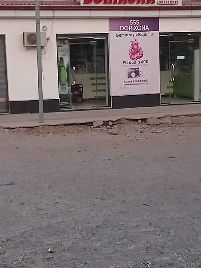 This is a vertical, slightly angled photograph taken outdoors in what appears to be a commercial area. The primary subject is a storefront with a large banner advertising a product called "DORIXONA 555 Flebodia 600." The banner is predominantly purple and white and features text in what appears to be Uzbek and Russian. The Uzbek text asks, "Gemorroy chiqdimi?" which translates to "Do you have hemorrhoids?" The Russian text asks, "Вылез геморрой?" which also translates to "Hemorrhoids have appeared?" and instructs to "Залей Слебодия 600," meaning "Pour Flebodia 600." The banner also contains an illustration of a pink, possibly floral or anatomical, element and a small image of a pill bottle.
To the left of the banner, a vertical sign with the number "95 109 97 77" is visible, likely a phone number. Behind the banner, the storefront windows reveal the interior of a pharmacy or medical supply store, with shelves and products visible. A small air conditioning unit is mounted on the exterior wall below the window.
The ground in front of the store is a rough, dusty surface with some scattered rocks and debris, suggesting a slightly unkempt urban environment. The overall impression is a daytime scene, likely in a town or city in Uzbekistan, given the language on the banner. There are no people visible in the frame.