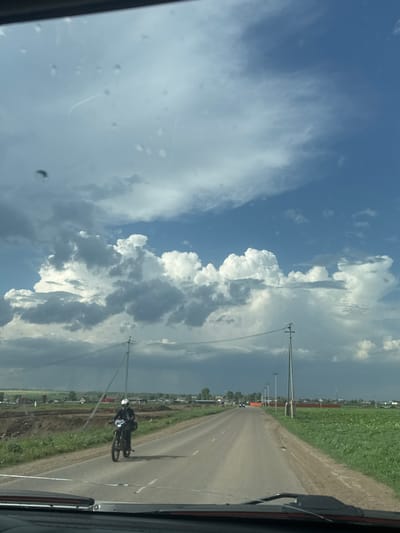 The image shows a view from inside a car, looking down a road in Kitsjanovo, Russia. A motorcyclist is traveling on the road in the same direction as the car. The sky is visible, with significant cloud cover. There are power poles running alongside the road. Fields or grassy areas border the road on both sides. The scene appears to be a rural or semi-rural environment.