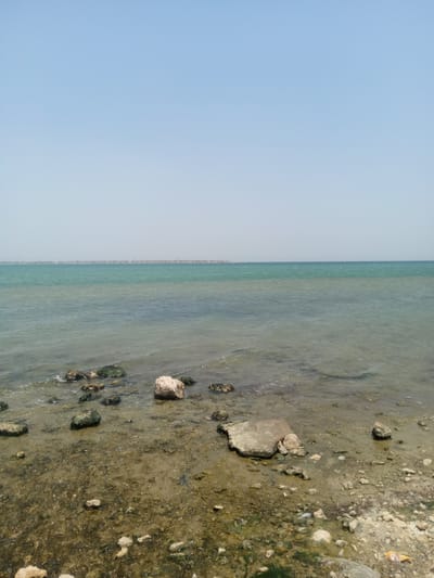 The image depicts a coastal scene in Al `Aziziyah, Saudi Arabia, characterized by a predominantly clear, pale blue sky that transitions to a hazy horizon. Below the horizon, a body of water extends towards the viewer, varying in color from a light turquoise in the distance to a more translucent, greenish-brown near the shore.

The shoreline is comprised of a mix of wet sand, small rocks, and larger boulders scattered throughout. There are traces of organic material mixed with the sand and rocks, suggestive of algae or vegetation. The foreground shows a close-up view of the tidal zone where the water meets the land, revealing an uneven surface covered with rocks, sand, and possibly some debris. The scene appears uninhabited and still.