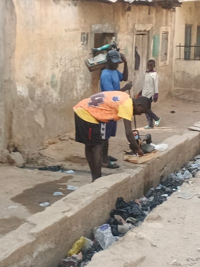 This image depicts an outdoor scene in Kwachiri, Nigeria, characterized by an unpaved ground surface and weathered buildings. In the foreground, a dark-skinned male, wearing an orange and yellow t-shirt with a blue number "4" on the back and dark shorts, is bending over. He is barefoot and attending to a small wooden crate positioned on the edge of a concrete-lined drainage ditch. The crate contains at least one dark-feathered fowl, possibly a duck, and several white objects on a white surface. The drainage ditch itself is filled with an accumulation of refuse, including numerous black plastic bags, lighter plastic debris, and other general litter.
Behind the bending individual, slightly to the left, stands another dark-skinned male, wearing a blue t-shirt and a dark baseball cap. He holds a dark stick in his right hand and carries a wooden frame, possibly for carrying goods or tools, on his left shoulder; a light blue object resembling a pipe or tool handle is visible on this frame. He wears dark sandals. Further in the background, a young child wearing a light-colored long-sleeved shirt, light-colored pants, and bright green shoes walks from left to right, looking towards the right side of the frame. The background consists of dilapidated, light brown or tan buildings with unpainted, weathered surfaces and simple window openings, some with wooden shutters. The ground near the buildings shows patches of moisture or standing water and additional litter, including white plastic bags. The overall lighting suggests daytime.