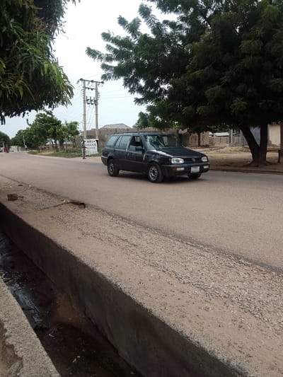 The image shows a black Volkswagen Golf station wagon driving on a paved road in Biliri, Nigeria. The car is traveling away from the camera. The windshield of the car is damaged. A concrete ditch is in the foreground on the left side of the road. The right side of the road has a tree. An electrical pole is along the left side of the road, with a sign that says "MAI ANGAMJE PALACE." A person stands near the sign. Buildings are visible further down the road. There are other trees in the background and foreground of the image.