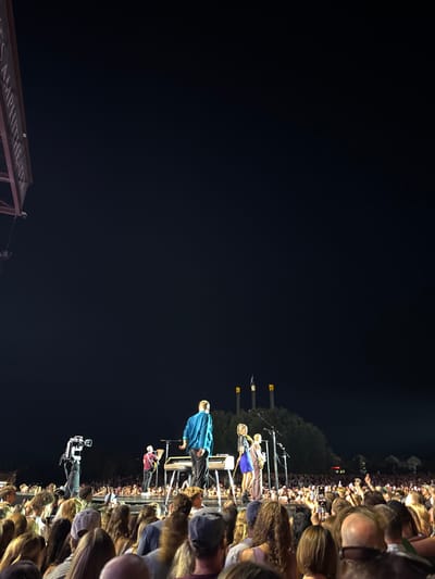 This image depicts a live outdoor concert at night, identified as the Les Schwab Amphitheater in Bend, Oregon, United States, indicated by the partial venue name visible in the upper left and the distinct three illuminated industrial stacks in the background, characteristic of the Old Mill District.
On the illuminated stage, a male performer in a blue shirt is positioned at a keyboard, facing away from the camera. To his right, two female performers stand near microphones; one is dressed in a blue skirt, and the other in a brown top. Further to the left on the stage, another male performer with a guitar is visible. A large video camera on a tripod is positioned on the far left of the stage. The foreground and midground are filled with a densely packed audience, primarily visible from behind, with many individuals holding up mobile phones to record or photograph the performance. The environment is dark, with the stage and some audience members illuminated by stage lights. Beyond the stage, a dark treeline is visible, topped by the three tall, light-colored structures. The sky overhead is dark and clear.