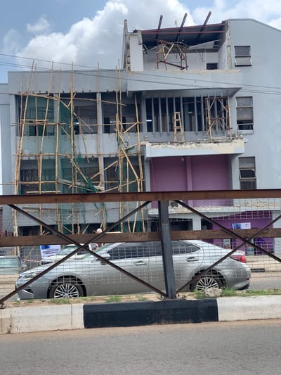 A silver car is parked on the side of a road in front of a building under construction. The building is located at the coordinates 7.252 latitude, 5.199 longitude, in the city of Ondo, Nigeria. The building is partially covered in scaffolding made of bamboo poles and netting. The roof is damaged, with pieces of the roof missing. There is a black and white curb next to the road, and a metal fence with a grid pattern. There is a sign in the lower left of the image that reads "INDEPENDENT ASSEMBLY." The building is painted in a pale gray color, and there are two windows on the upper level and two on the lower level. The ground appears to be made of asphalt and dirt. There are no people visible in the image. The sky is cloudy with scattered patches of blue.